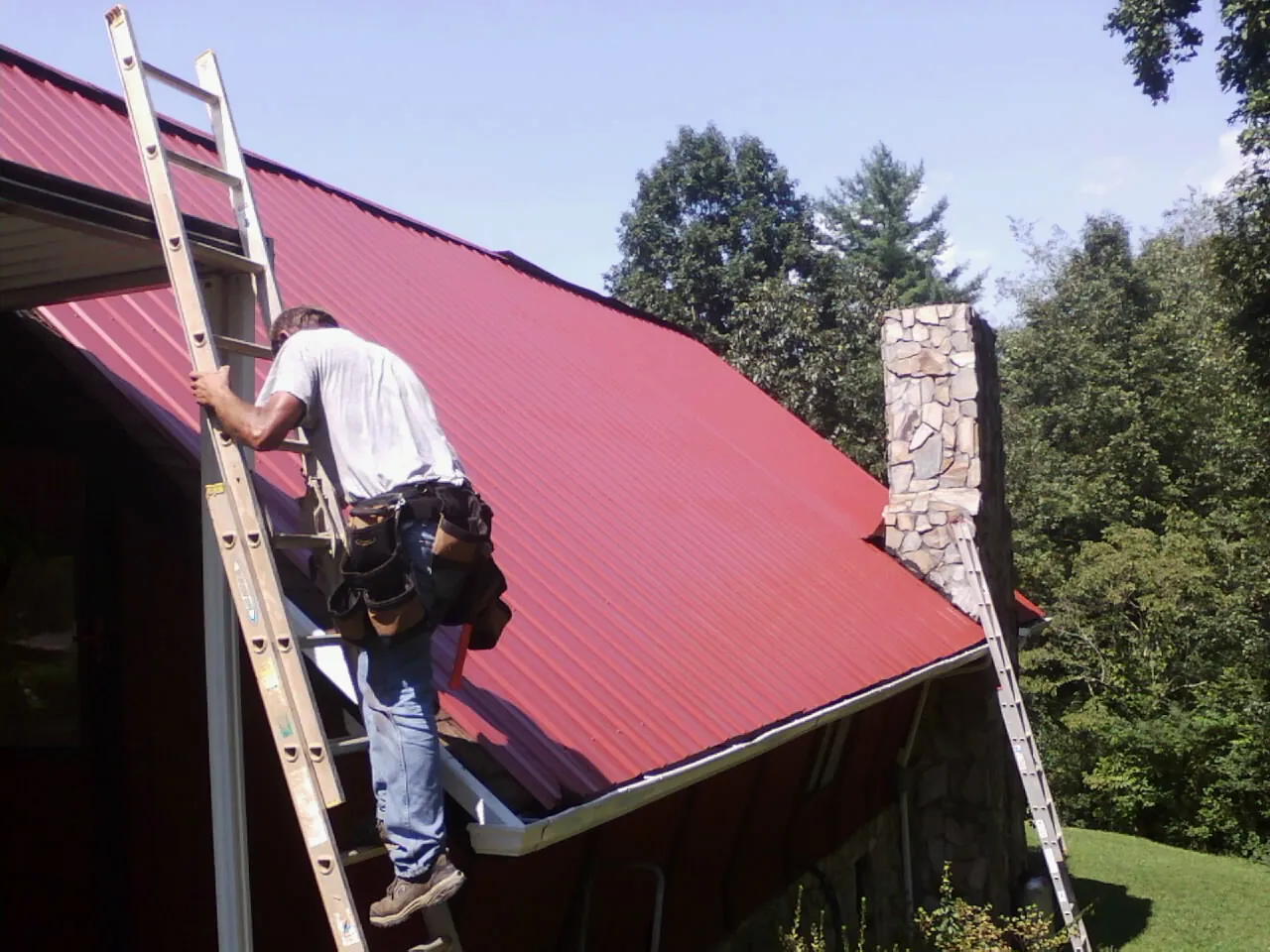 Roofer on a ladder beside a red metal roof and stone chimney — Soffit & Fascia Repair by Roofing Brothers in Gates Mills
